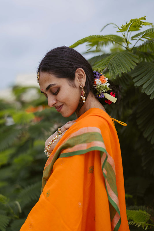 actress in an orange jamdani saree with floral hairpiece standing among green foliage