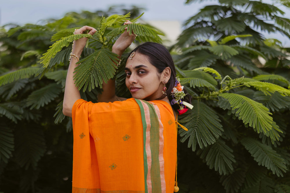 model in an orange traditional jamdani saree outfit standing among green foliage