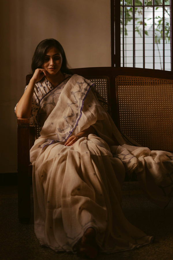 A woman sitting on a chair, draped in a beige and indigo handloom linen jamdani saree with a running blouse.