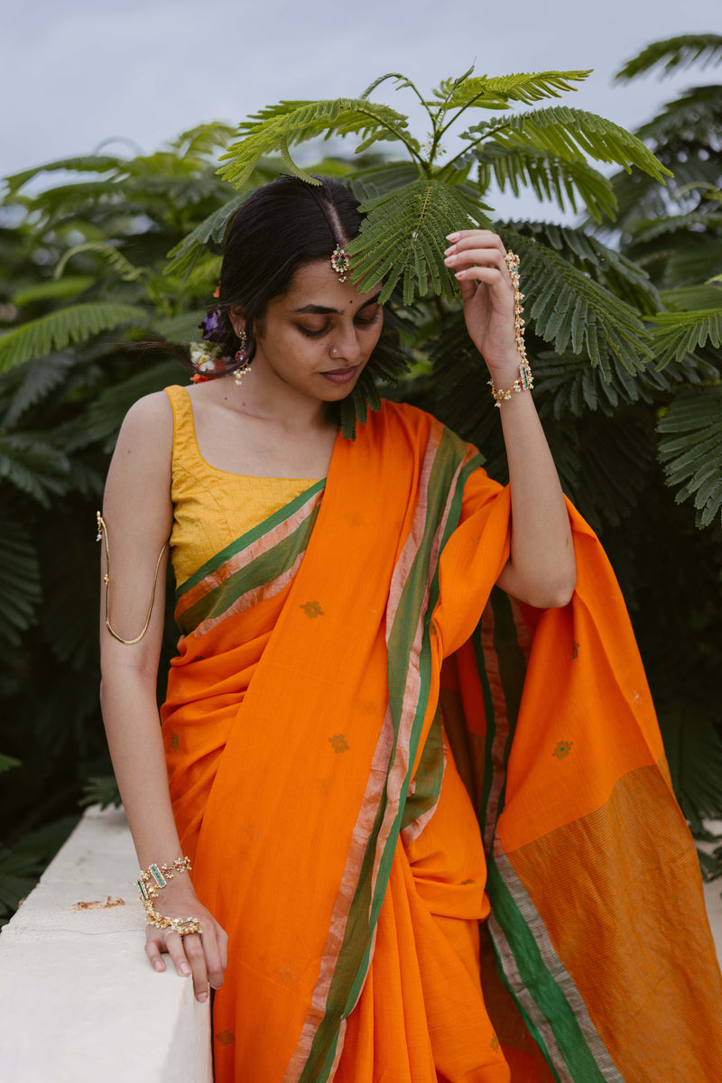 Elegant handloom cotton saree with orange body, green border, and intricate zari motifs.
