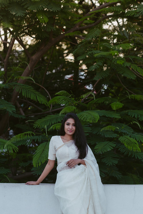 Woman in a white LINEN SILK saree standing against a backdrop of green foliage