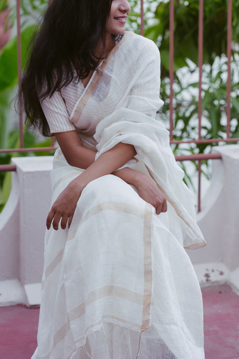 Woman in a white saree sitting outdoors with greenery in the background