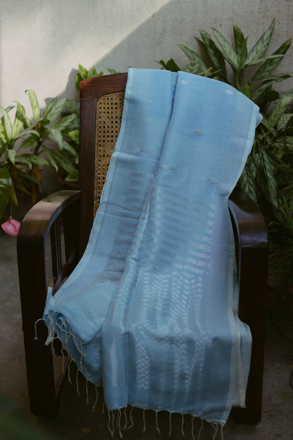 Baby blue handloom linen Jamdani saree with silver motifs displayed on a wooden chair