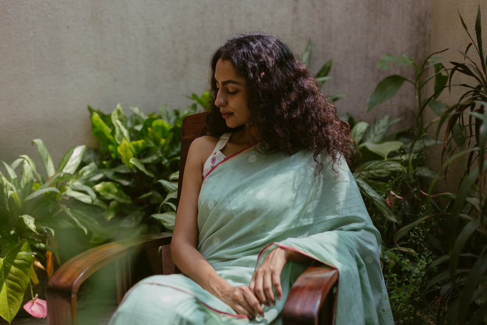 Woman in a light green jamdani cotton saree sitting in a garden with plants around