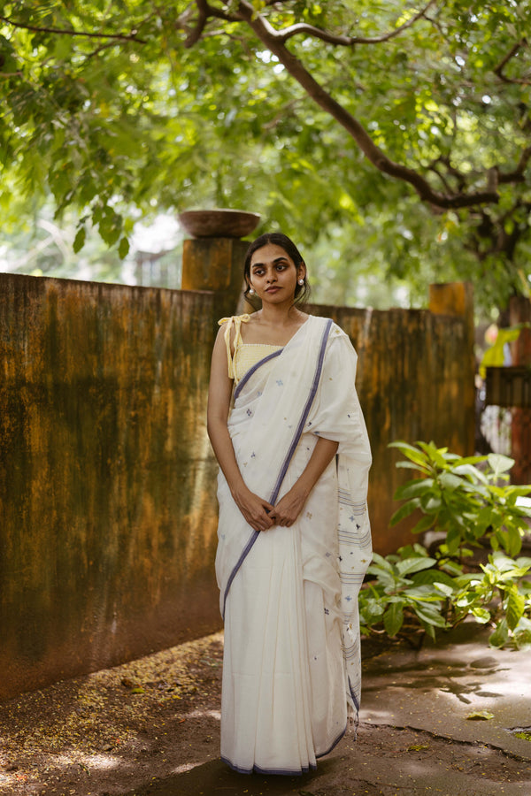 Model wearing a white handloom cotton indigo jamdani saree with floral motifs on the pallu, styled elegantly with a chic yellow bow blouse.
